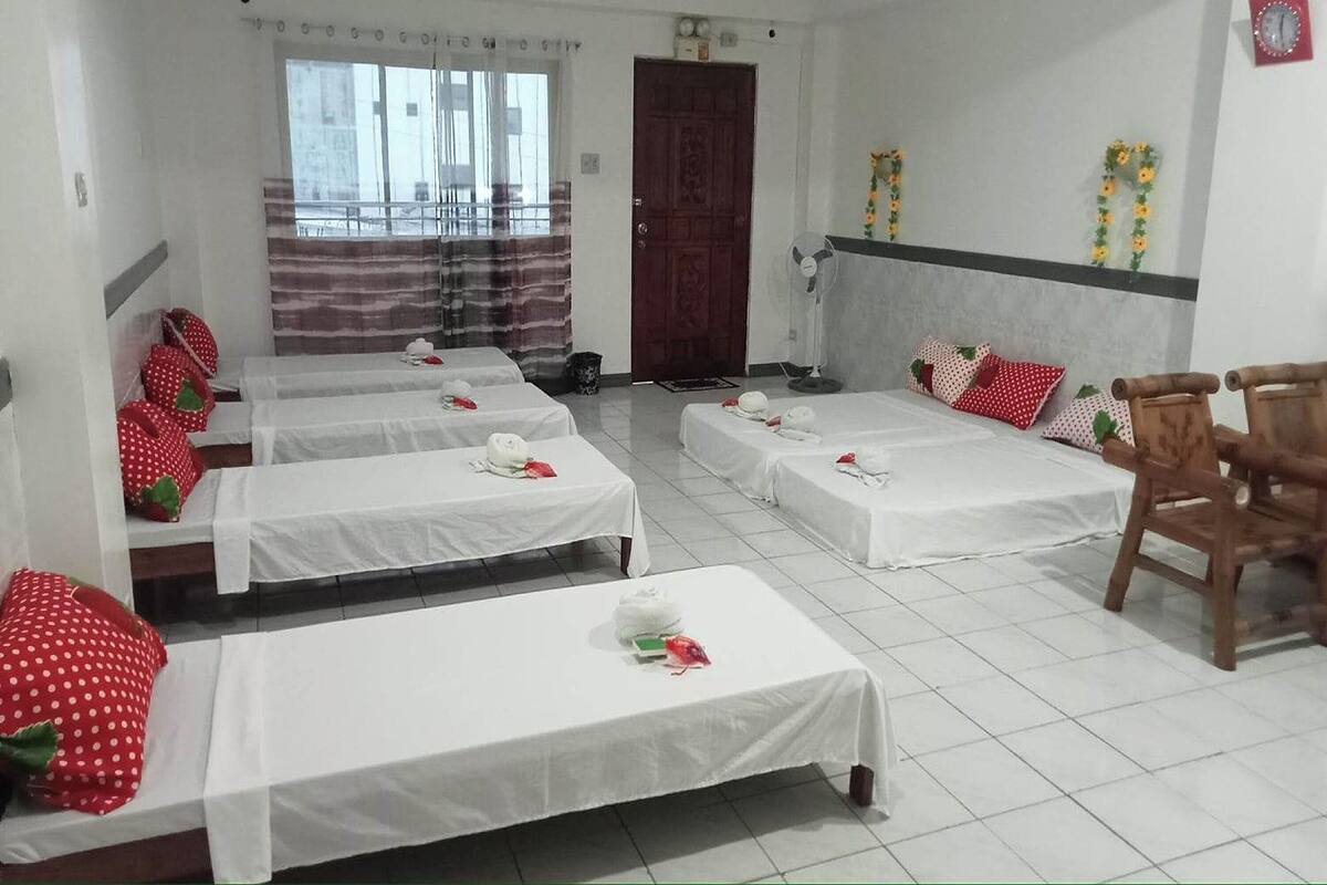 A hotel room with four single beds, each with a white bedspread and red pillows. There is a wooden chair and a small table with a white tablecloth. The room has a balcony with a striped curtain and a view of buildings outside.