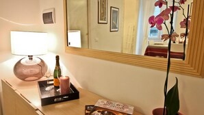 Een hotelkamer met een houten bureau, een spiegel, een lamp, een dienblad met een fles en kaars, een boek getiteld 'Tuscany Interiors', en een potplant.