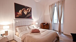 Een slaapkamer met een groot bed, een hoofdeinde, een nachtkastje, een lamp, een schilderij en een raam met gordijnen.