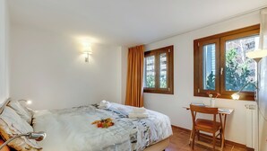 Une chambre à coucher comprenant un lit, un bureau, une chaise, une lampe et deux fenêtres avec des rideaux.