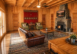 Interior de una cabaña de troncos con chimenea, un sofá de cuero, dos sillones y una alfombra estampada.