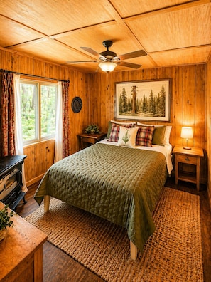 Ein Schlafzimmer einer Holzhütte mit Bett, Deckenventilator, Fenster mit Vorhängen, einem Gemälde, einem Nachttisch und einer Lampe.