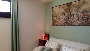 A bedroom with a bed, pillows, a nightstand with a lamp, a painting on the wall, and a window with curtains. - Verde Mare apartment in villa with garden, oasis Saline and Playa Sol Taguali (Carrozziere)