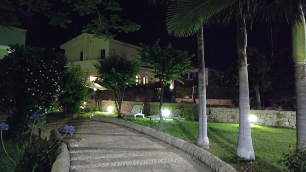 A nighttime view of a paved pathway leading to a building with a stone wall and a palm tree. - Verde Mare apartment in villa with garden, oasis Saline and Playa Sol Taguali (Carrozziere)