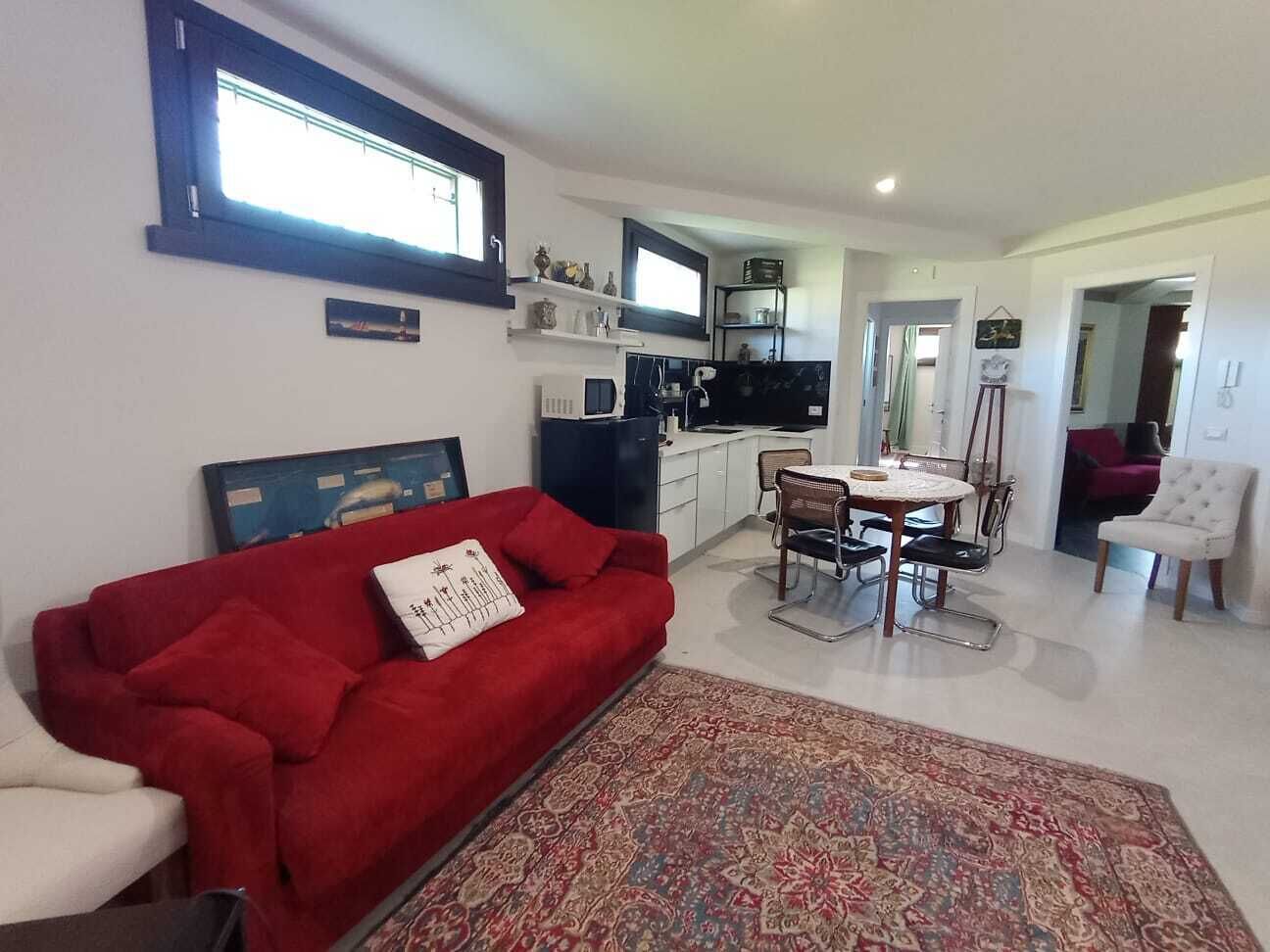 A living room with a red sofa, a dining table with chairs, a kitchen area, and a small white chair.