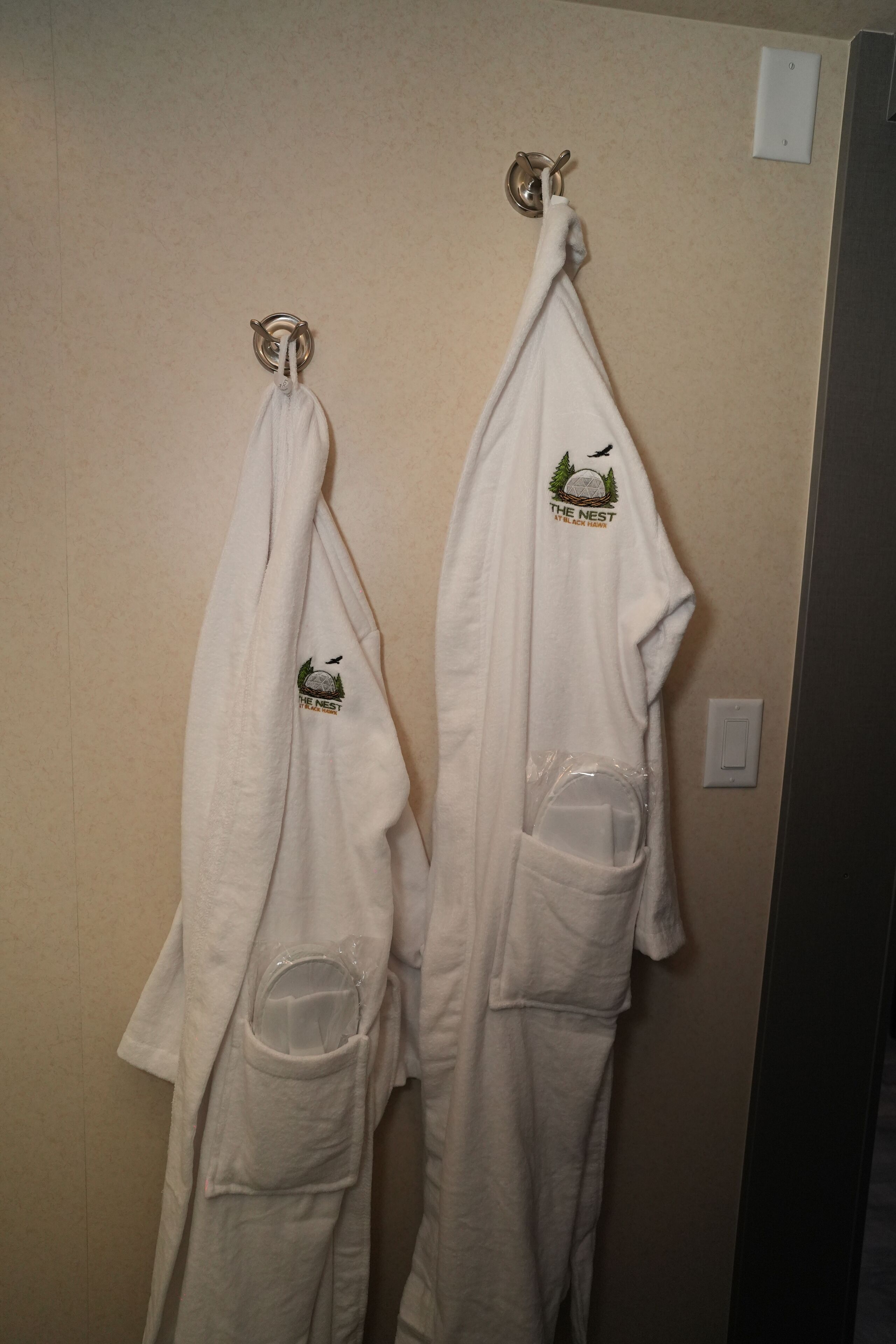 Two white robes hanging on hooks against a beige wall, each with a logo and a pocket.