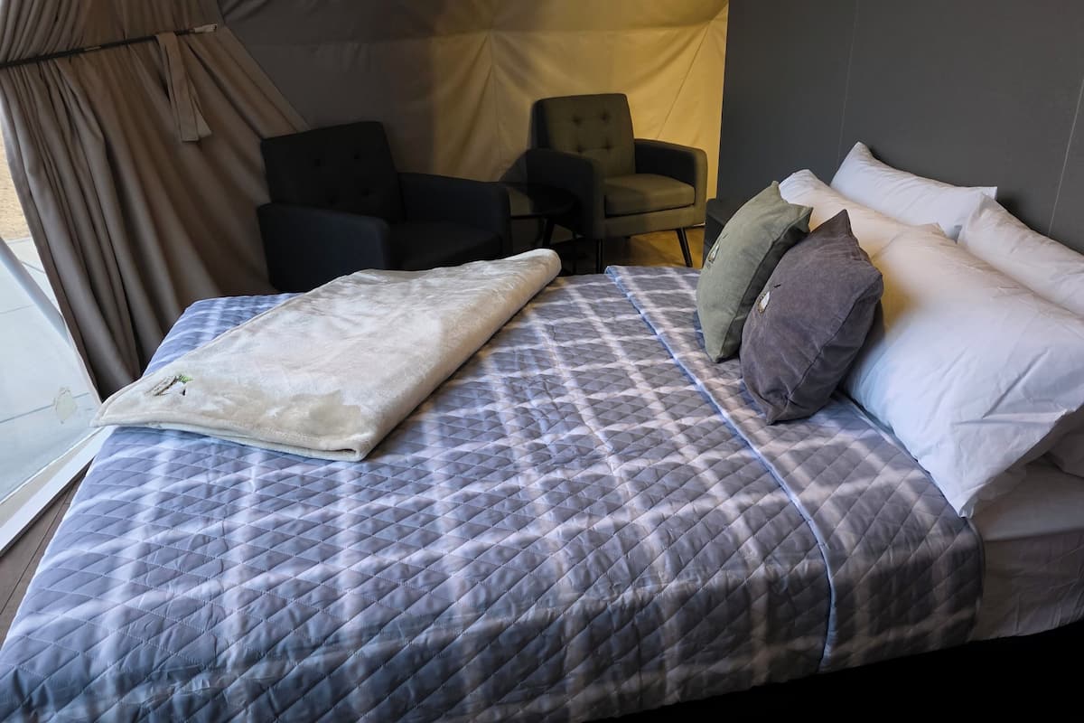 A neatly made bed with a quilted cover, pillows, and a blanket. There is a chair and a small table in the room.