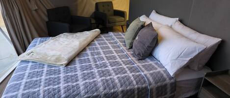 A neatly made bed with a quilted cover, pillows, and a blanket. There is a chair and a small table in the room.