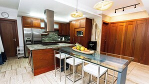 Une cuisine moderne avec des meubles en bois, des appareils en acier inoxydable, un îlot central en verre avec des tabourets de bar et un luminaire suspendu.