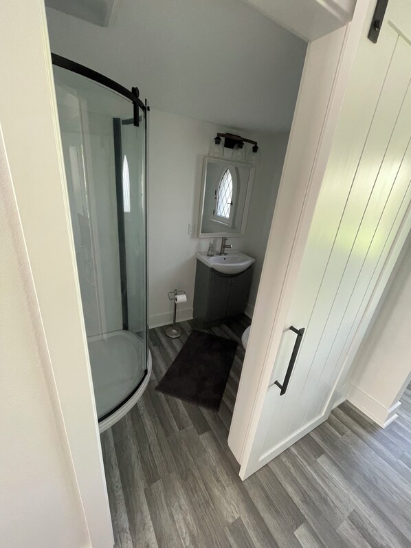 Un baño pequeño con cabina de ducha de vidrio, lavamanos y una ventana sobre el espejo.