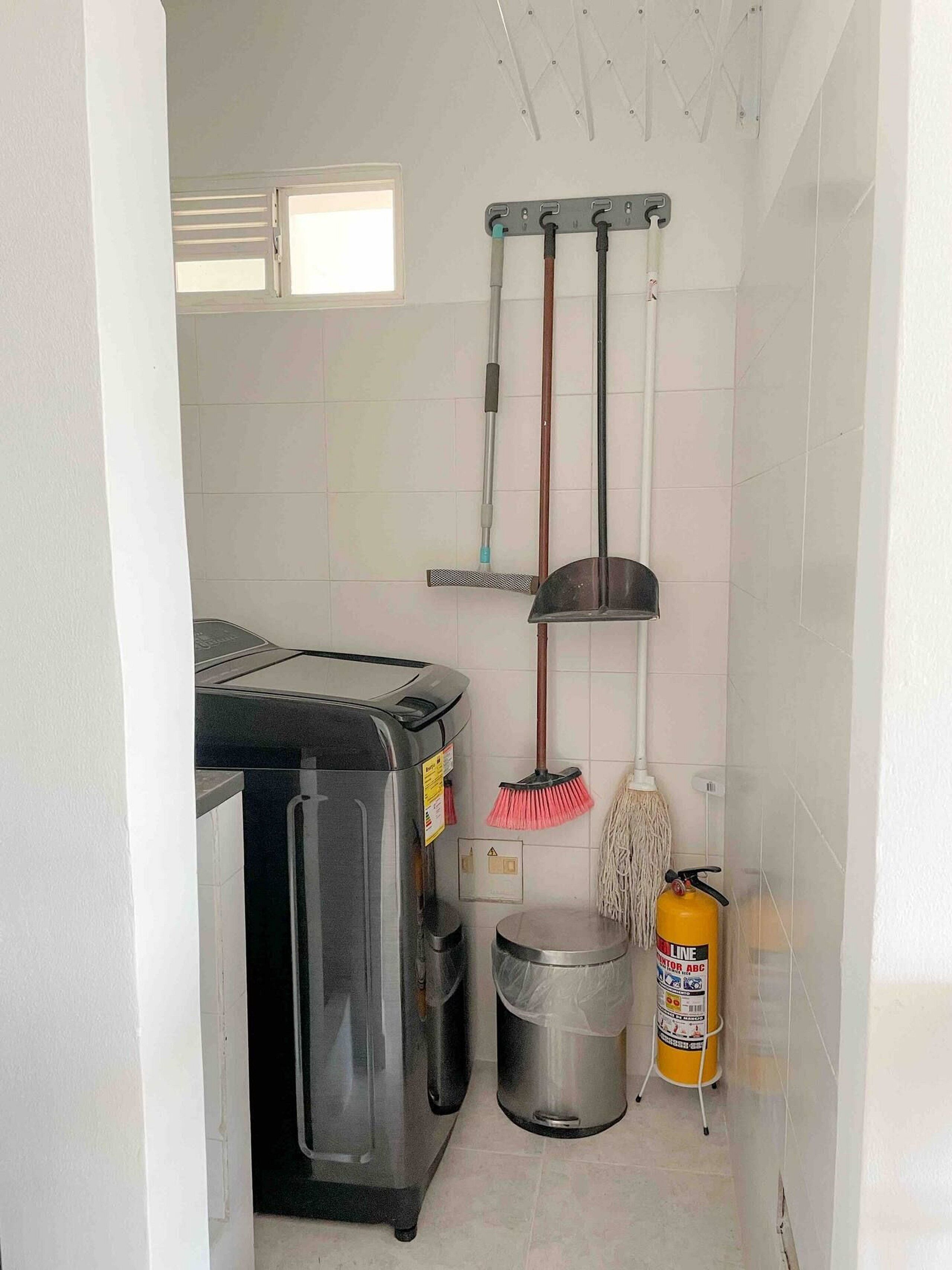 Eine kleine Küche mit einem Kühlschrank, einem Mülleimer, einem Mop, einem Besen und einem Feuerlöscher.