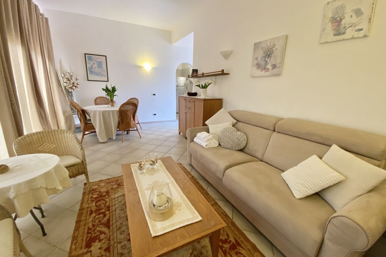 Ein Wohnzimmer mit einem beigen Sofa, einem runden Esstisch mit Stühlen, einem Couchtisch mit Glas und Kerze sowie einer kleinen Kommode mit Dekoration.