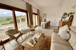 A spacious living area with a sofa, a coffee table, and a dining table set with chairs. There is a balcony with a view of greenery.
