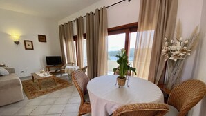 Ein Wohnzimmer mit rundem Tisch, Korbstühlen, einer Couch, einem Fernseher und Blick ins Freie.