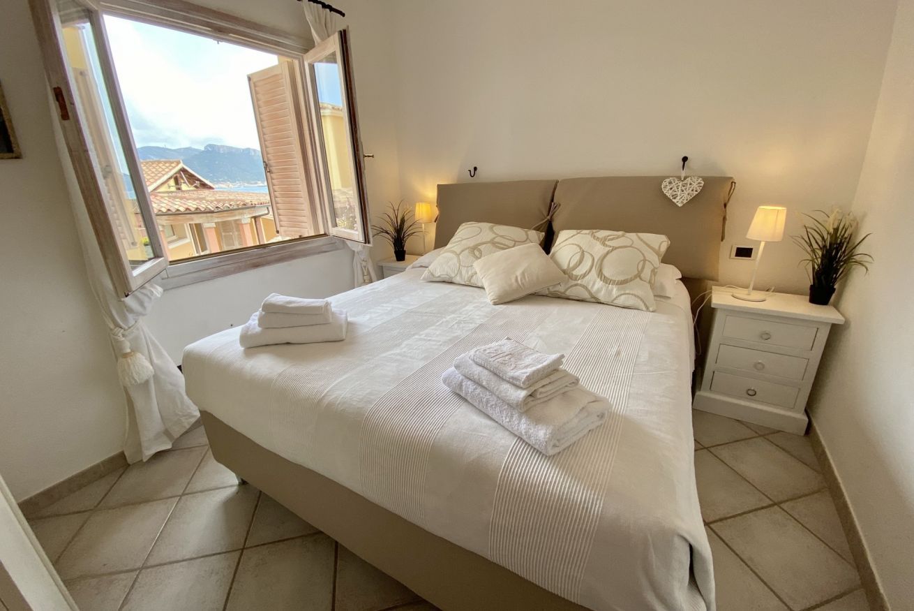 Un lit bien fait, avec du linge de lit blanc et des serviettes pliées, une table de chevet avec une lampe, et une vue sur les bâtiments par la fenêtre.