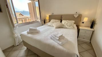 Un lit bien fait, avec du linge de lit blanc et des serviettes pliées, une table de chevet avec une lampe, et une vue sur les bâtiments par la fenêtre.