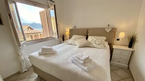 Ein ordentlich bezogenes Bett mit weißen Leinentüchern und gefalteten Handtüchern, ein Nachttisch mit Lampe sowie ein Blick auf Gebäude durch ein Fenster.