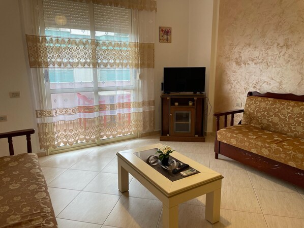 Una sala de estar con un sofá, una mesa de centro, un televisor sobre un soporte de madera y una ventana con cortinas de encaje.