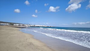 Une plage avec un ciel dégagé et des vagues. On aperçoit des bâtiments à l’arrière-plan.