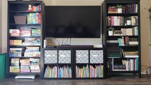 A living room with a large flat-screen TV, two black bookshelves filled with books and games, and a small table with a white vase and a plant. - Charming 4-bedroom home, High Speed Internet & AC in beautiful Colorado Springs (Colorado Springs)