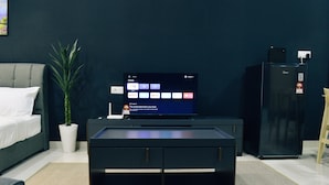 Una sala moderna con un televisor de pantalla plana sobre un pedestal negro, un sofá gris, una planta en maceta blanca y un refrigerador negro.