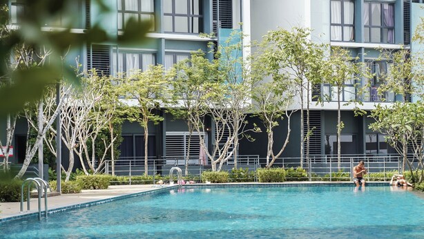 Una piscina con bordes de mosaico, rodeada de árboles y un edificio moderno con múltiples ventanas y balcones.