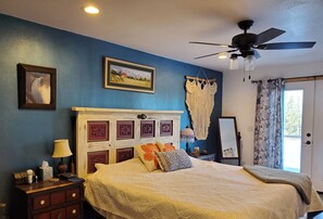 Une chambre à coucher comprenant un lit, une table de chevet, une lampe, des tableaux encadrés, une décoration murale en macramé et un ventilateur de plafond.