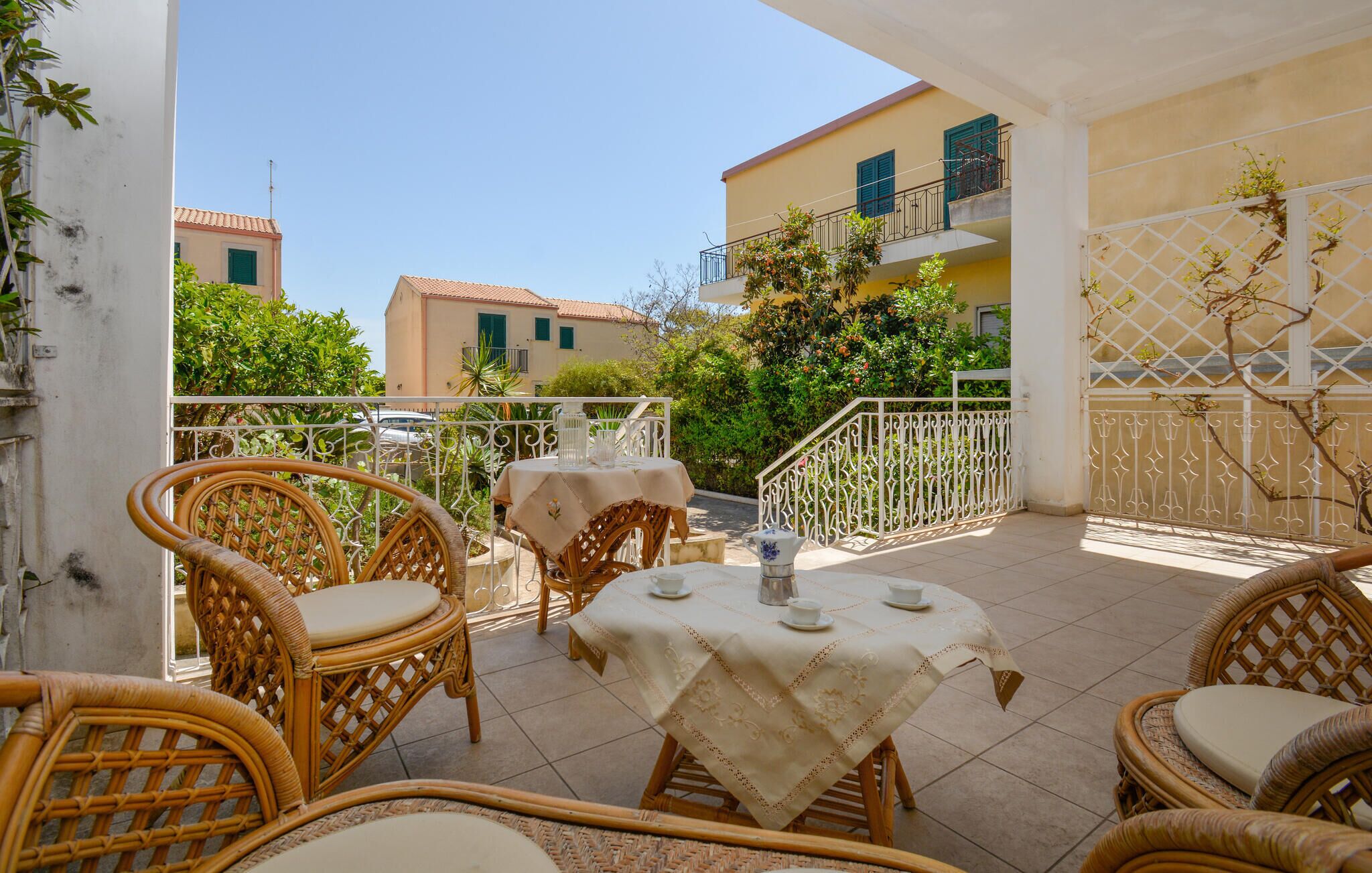 An outdoor patio with wicker furniture and a table set for two, surrounded by a white fence and greenery.