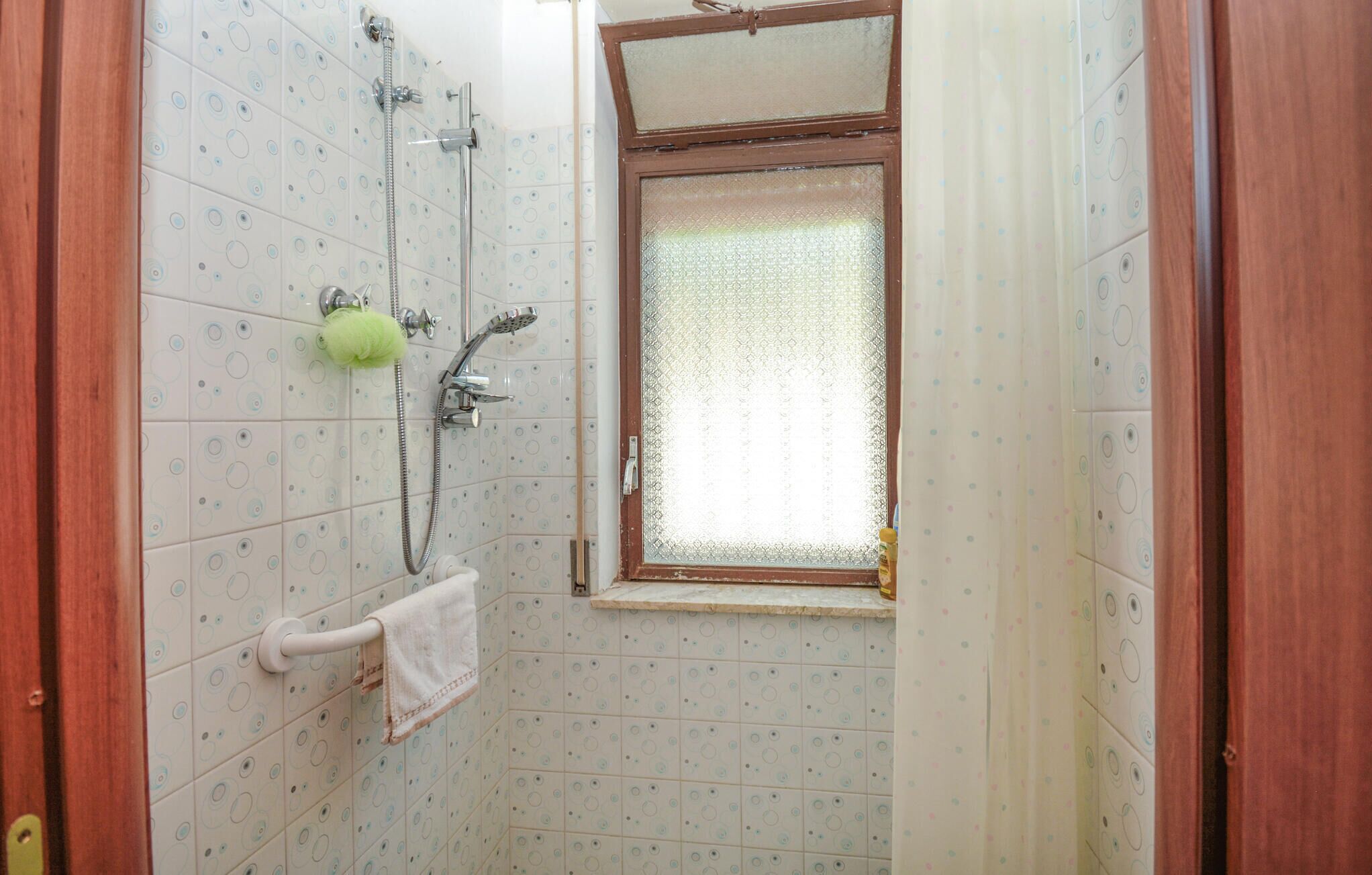 Een badkamer met een douche, een raam met gordijn, en een handdoek aan de muur.