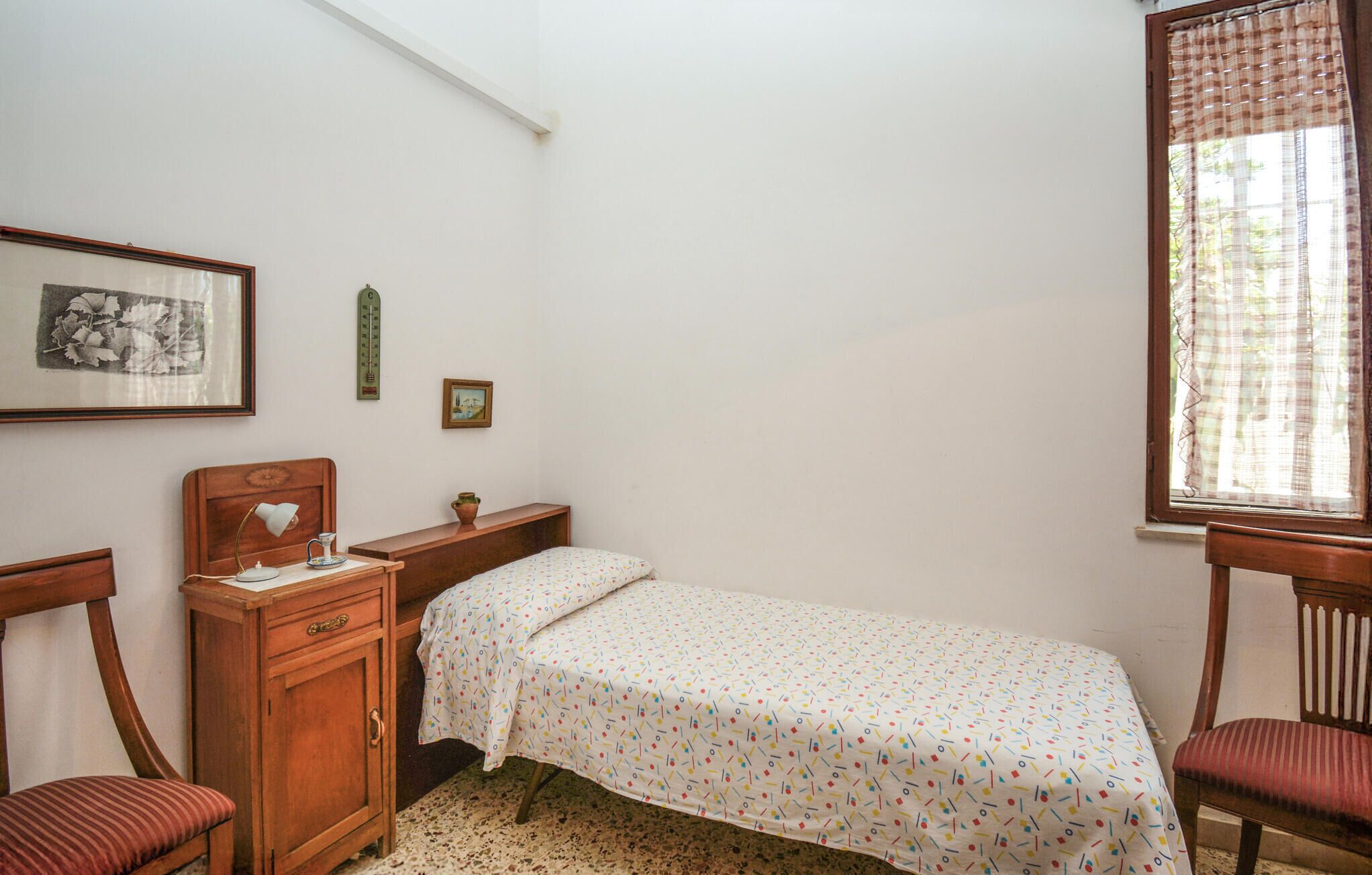 A small, simple bedroom with a single bed, a nightstand, a wooden dresser, two chairs, and a window with curtains.