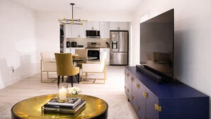 Una sala de estar moderna con un televisor de pantalla plana, una mesa de centro con libros, un comedor con sillas y una cocina al fondo.