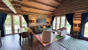 Interior acogedor de una cabaña con techo de madera, una mesa de comedor con sillas, un sofá verde, un tapete estampado y vistas al follaje exterior.