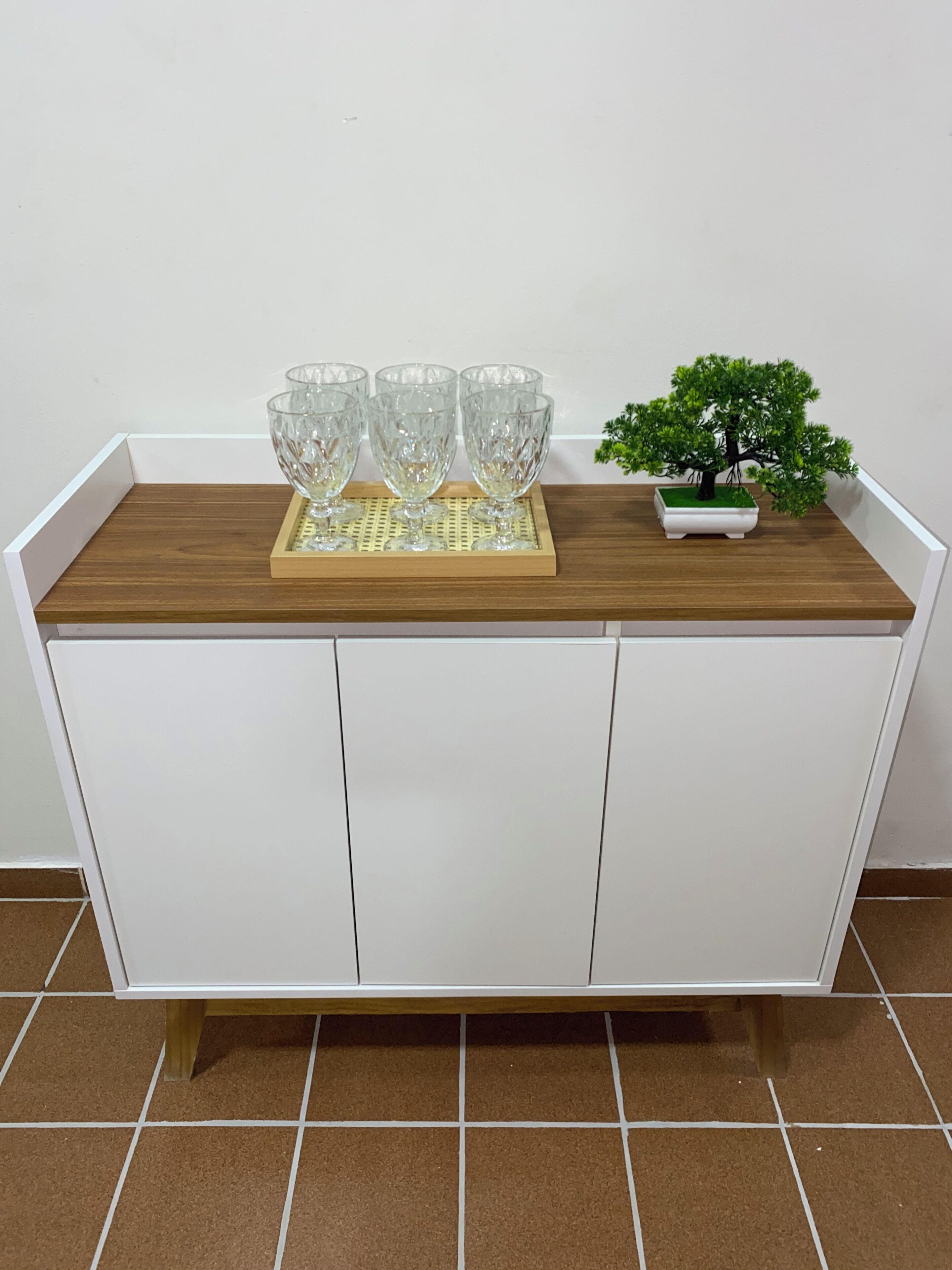 A white cabinet with a wooden top shelf, three clear glass cups, and a small potted plant.