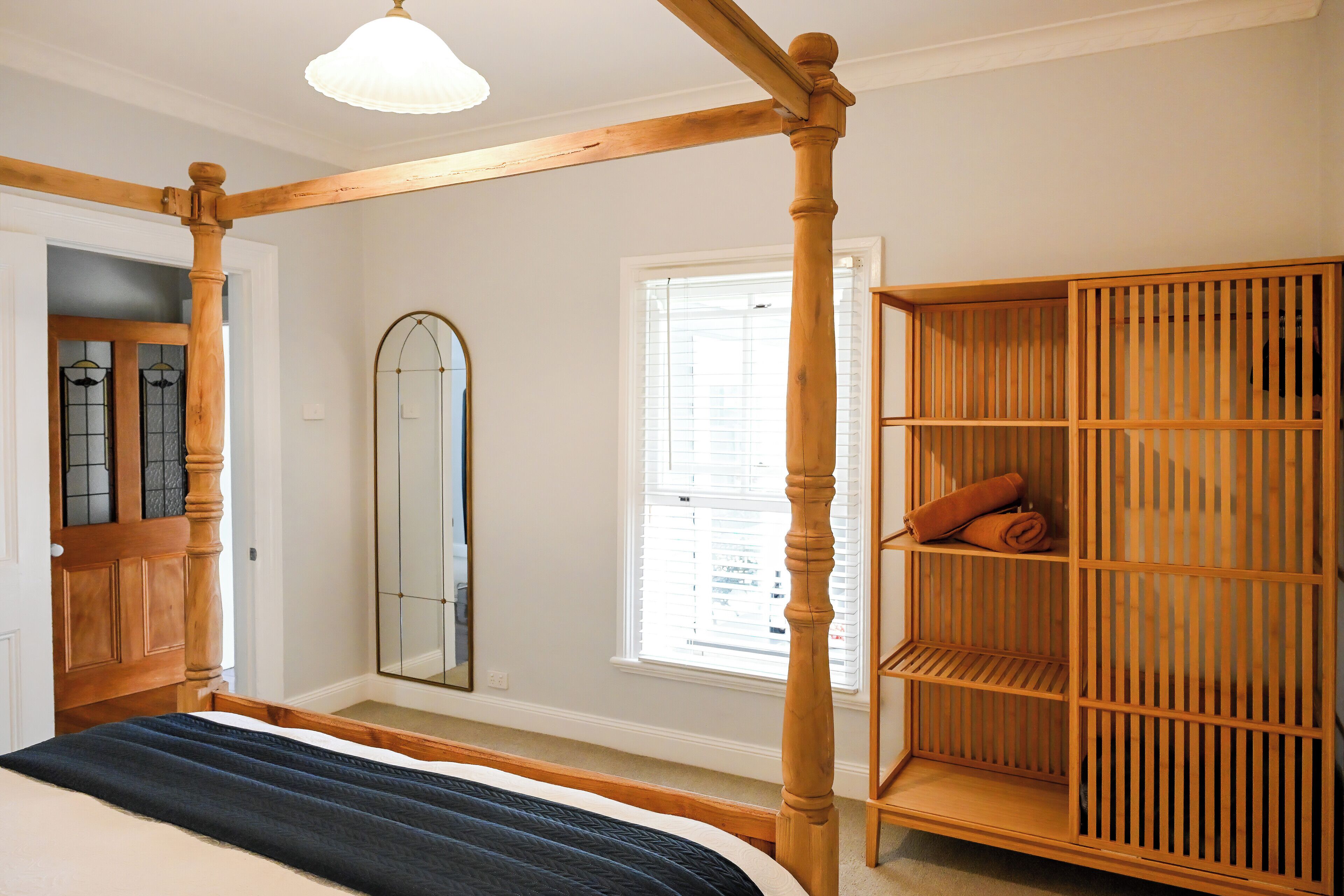 Une chambre à coucher avec un lit à baldaquin, une armoire en bois, un grand miroir et une fenêtre avec des stores.