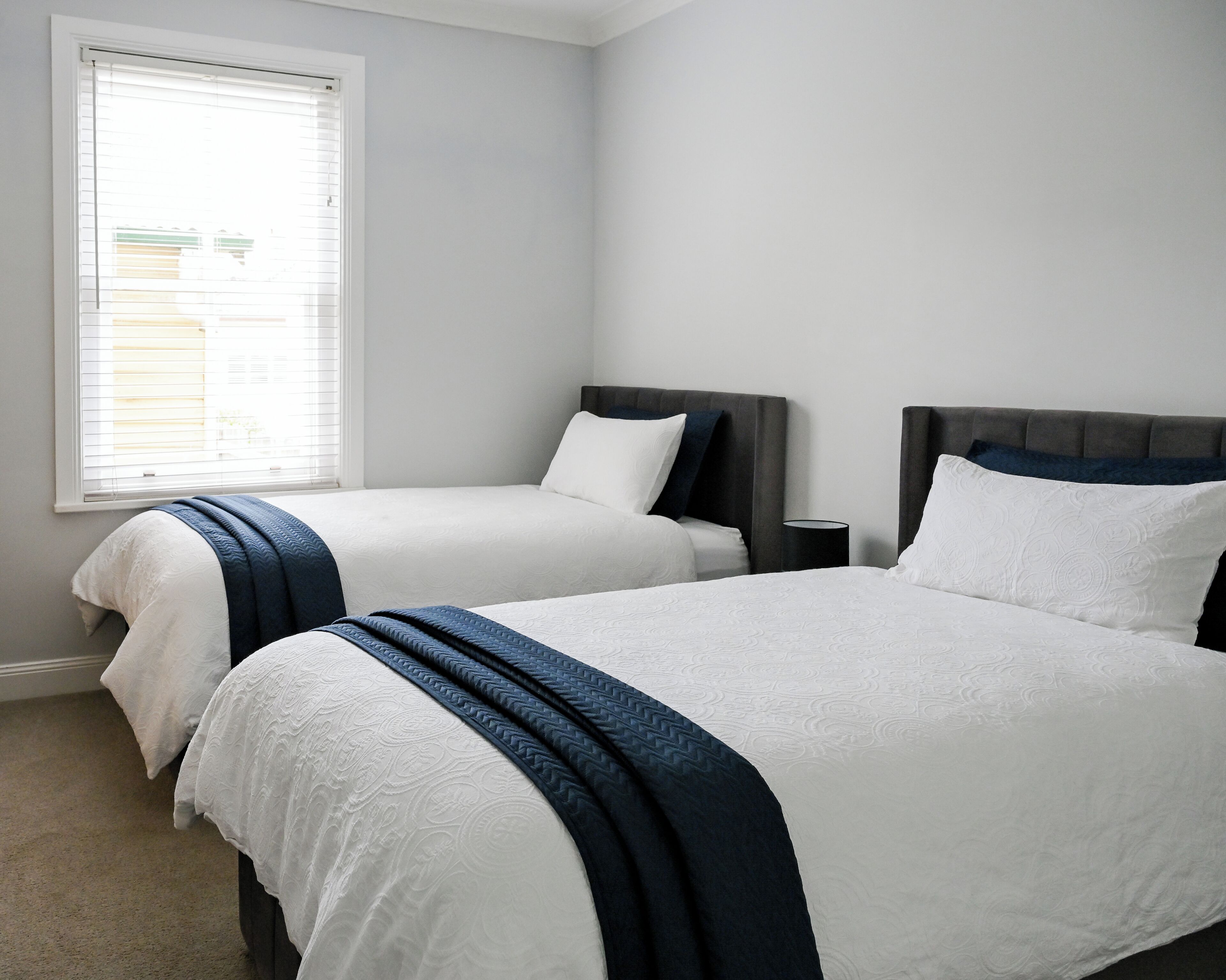 Ein Zimmer mit zwei Betten, einem Fenster mit Jalousien und einem Nachttisch mit Lampe.