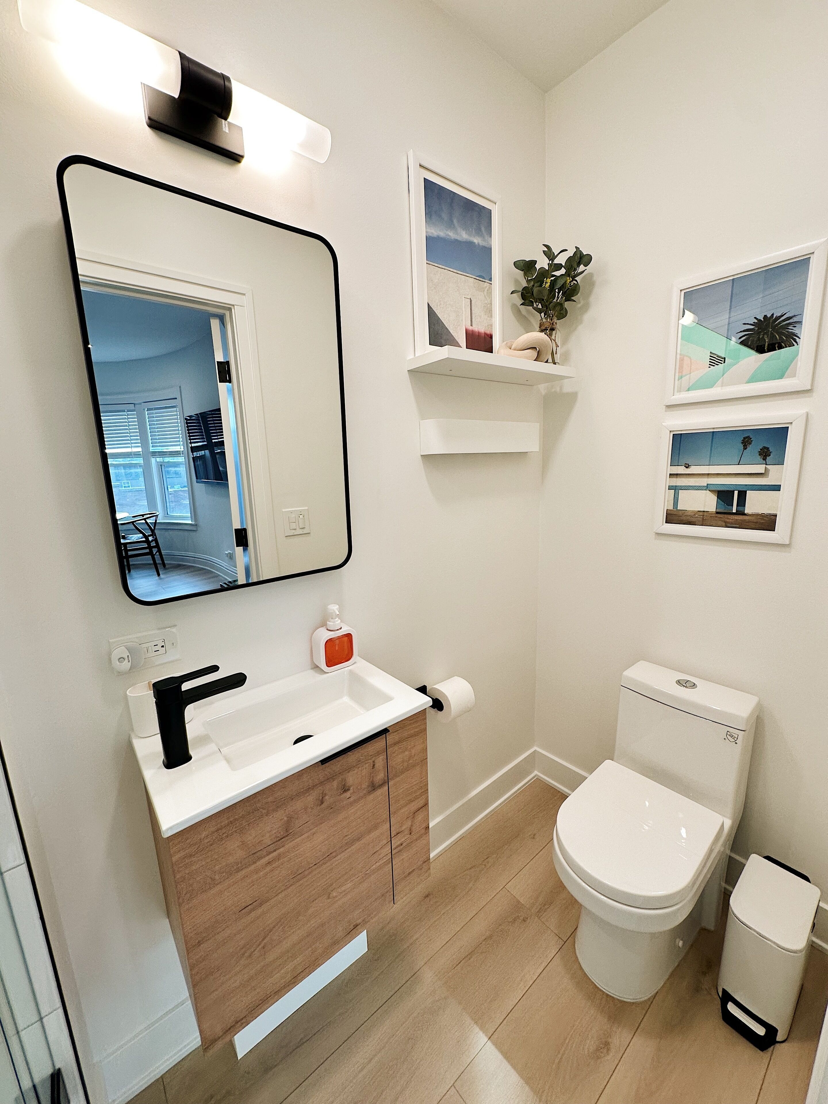 A modern bathroom with a white sink, black faucet, and a toilet. There are two framed pictures on the wall, a small plant on a shelf, and a mirror above the sink.
