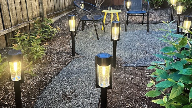 An outdoor patio area with a wooden fence, outdoor seating with chairs and a table, and multiple ground lamps.