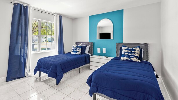 Une chambre avec deux lits, une fenêtre avec des rideaux, un miroir et un mur d’accent bleu.