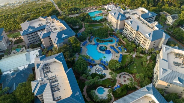 An aerial view of a resort with multiple buildings, a central pool area, and a beach in the background.