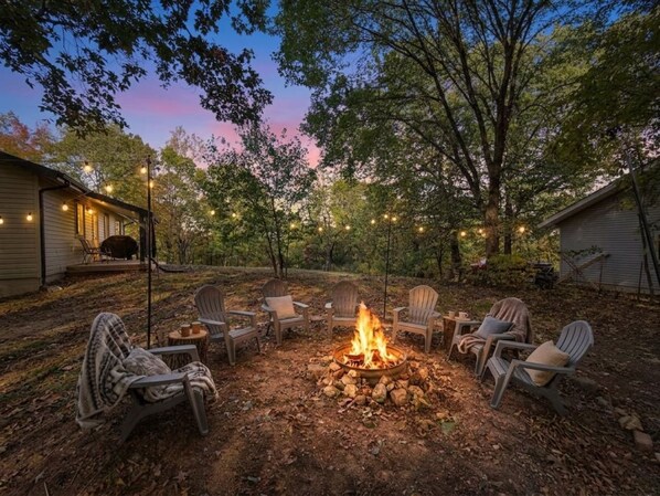 A fire pit surrounded by outdoor seating, with trees and a house in the background.