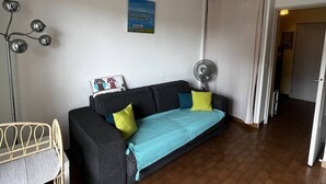 A living room with a sofa, a floor lamp, a painting, and a fan. - Studio with Cabin for 4 – Marina View, Beach Just Steps Away, with Elevator Access (CANET PLAGE)