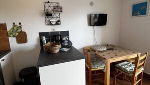 A compact kitchen with a dining area, a ceiling fan, a TV, and a small trash bin. - Studio with Cabin for 4 – Marina View, Beach Just Steps Away, with Elevator Access (CANET PLAGE)