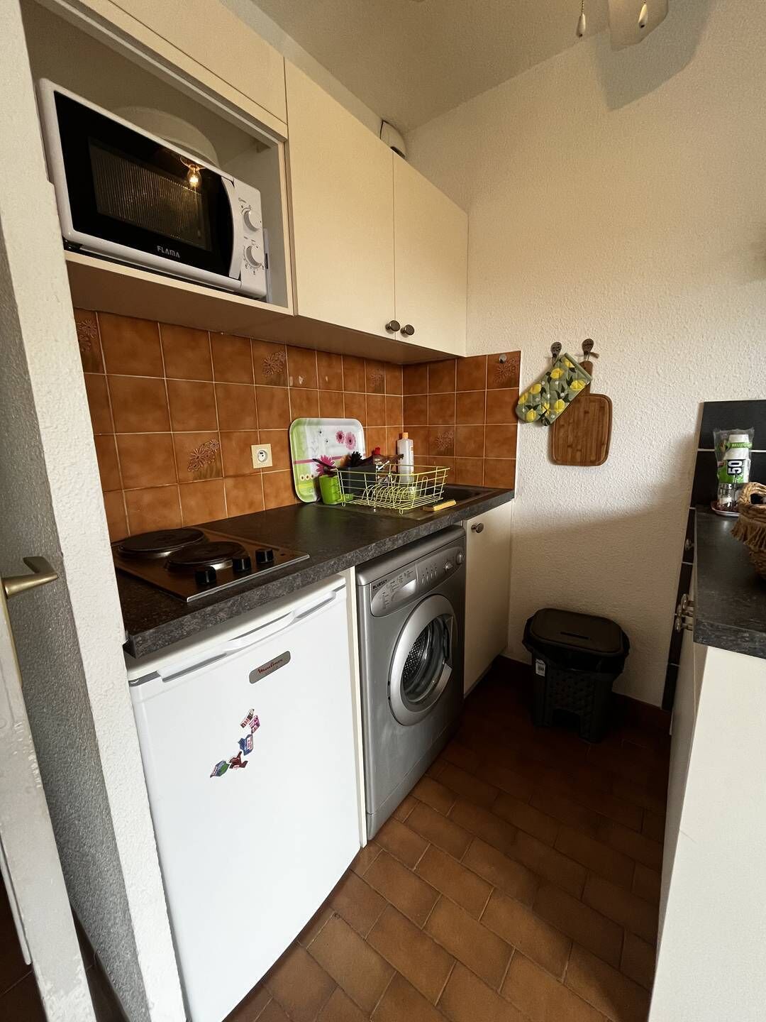 A compact kitchen with a microwave, a washing machine, and a small trash bin.