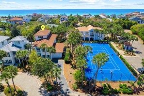 Vista aérea de un barrio residencial con una cancha de tenis rodeada de casas y palmeras.