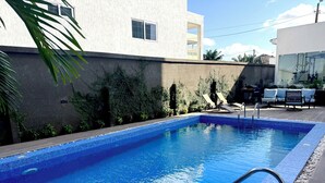 A rectangular swimming pool with a tiled edge, surrounded by a dark wall with greenery and a clear sky. - Luxury studio pool/gym in east legon at Belrose (Accra)