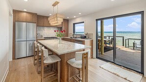 A modern kitchen with a large island, stainless steel appliances, and a view of the ocean through the sliding glass doors. - 🌅 NEW! Waterfront Retreat on West Bay – Hot Tub, Games & Zen Den ➠ 7345 (Traverse City)