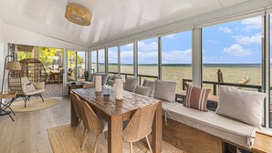 A spacious room with a wooden table and chairs, a sofa with cushions, and large windows offering a view of the water. - 🌅 NEW! Waterfront Retreat on West Bay – Hot Tub, Games & Zen Den ➠ 7345 (Traverse City)