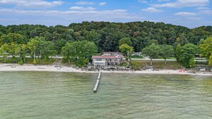 A waterfront property with a dock extending into a lake, surrounded by lush greenery and a hill in the background. - 🌅 NEW! Waterfront Retreat on West Bay – Hot Tub, Games & Zen Den ➠ 7345 (Traverse City)