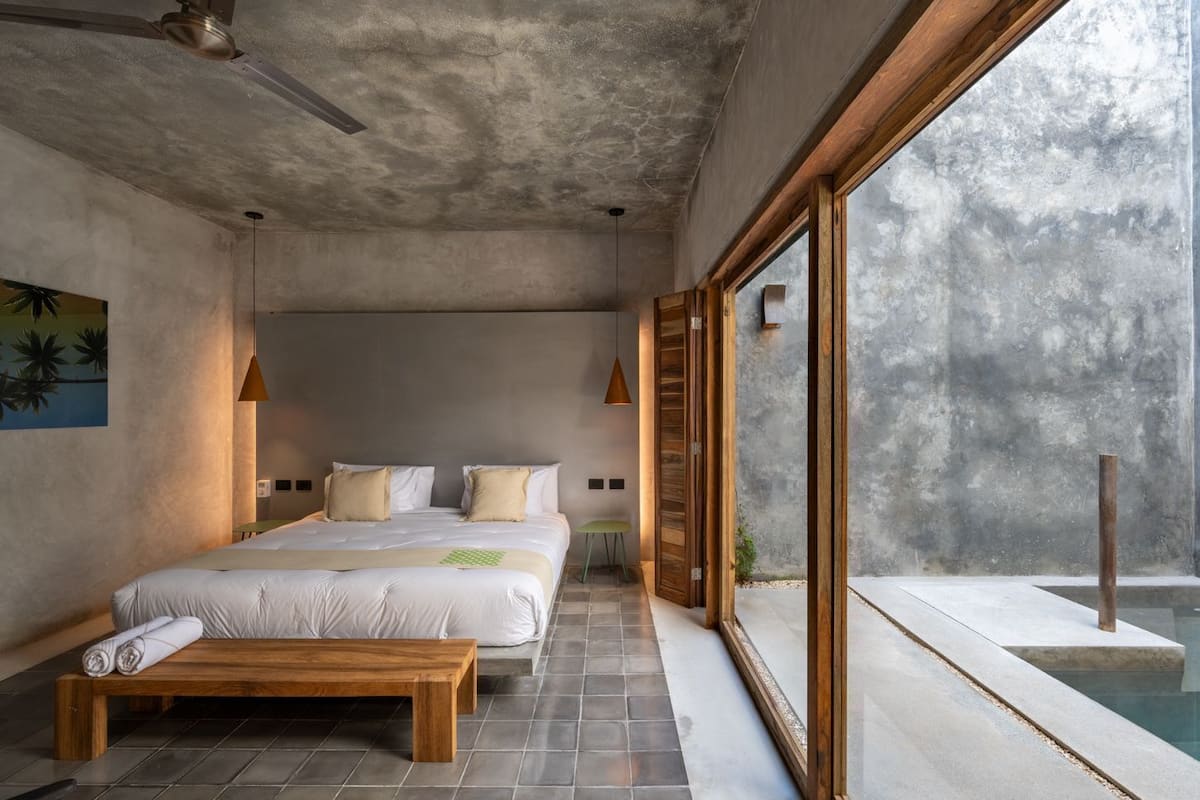 A modern bedroom with a large bed, a wooden bench, a view of a swimming pool, and a ceiling fan.