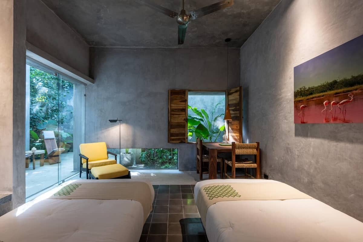 A modern room with two beds, a dining table, chairs, a ceiling fan, and a large window with a view of greenery.